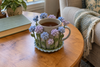 Blue and lilac crochet hydrangea heads and two butterflies surround a pale ceramic mug of tea on a round wooden table.