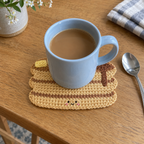 A pale blue coffee mug stands on a flat pancake stack crochet coaster with butter, syrup and a tiny smile on a breakfast table.