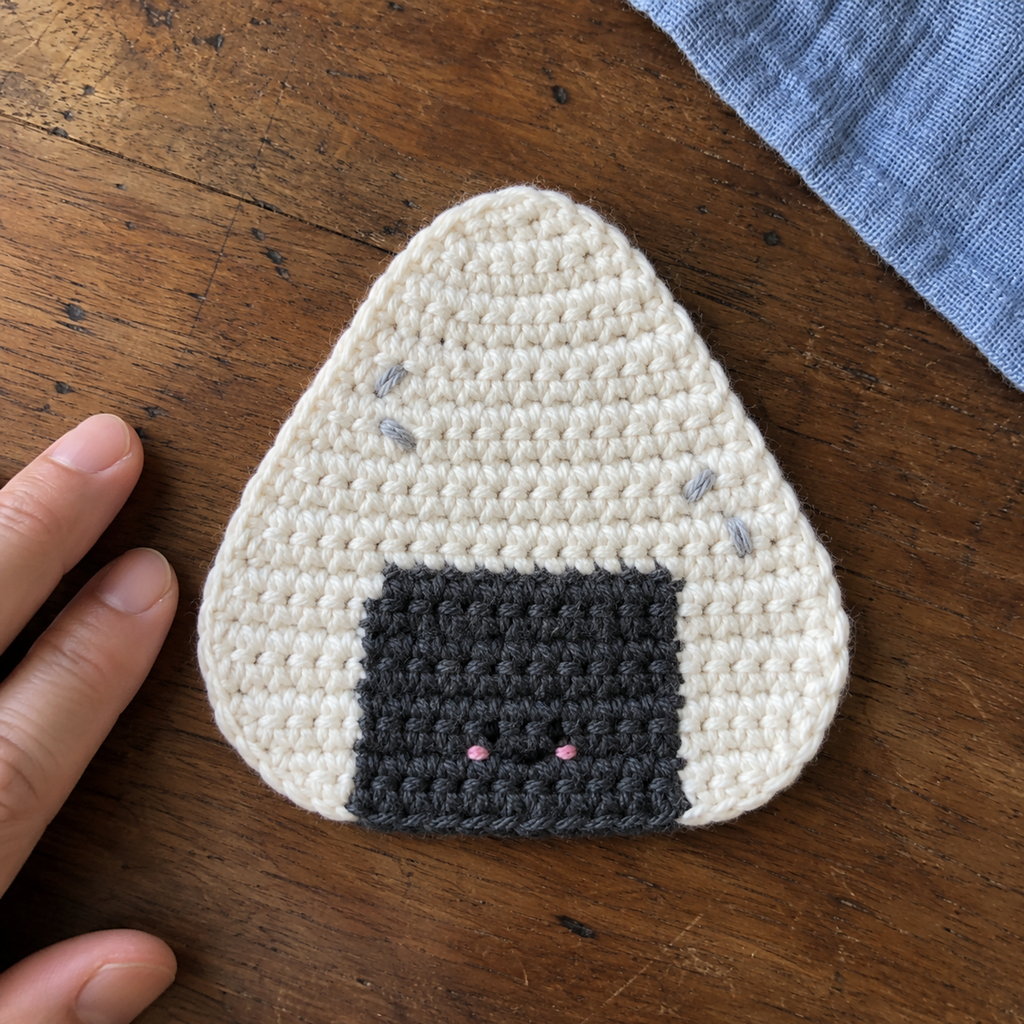 Close view of a white triangular onigiri crochet coaster with a dark nori patch and tiny embroidered face beside adult fingertips.