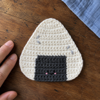 Close view of a white triangular onigiri crochet coaster with a dark nori patch and tiny embroidered face beside adult fingertips.