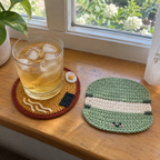 A glass of iced drink stands on a round ramen crochet coaster beside a green macaron coaster on a sunny wooden windowsill.
