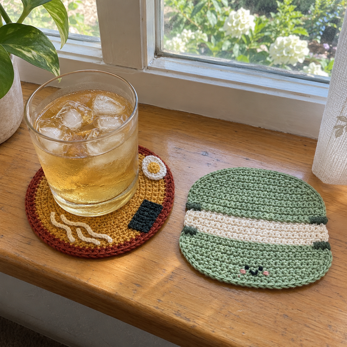 A glass of iced drink stands on a round ramen crochet coaster beside a green macaron coaster on a sunny wooden windowsill.