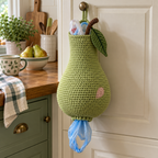 A green pear crochet bag holder with a low rounded bulb, pink cheek, leaf and curved brown stem hangs from a farmhouse cabinet knob.