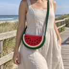 A small watermelon crochet crossbody bag worn over a linen summer dress on a seaside boardwalk, with red flesh, black yarn seeds and curved green rind bands.