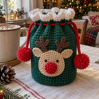 Angled detail of a green reindeer crochet pouch showing its tan face, branched brown antlers, raised red nose, white ruffled cuff, eyelets and red pompom-ended drawstring.