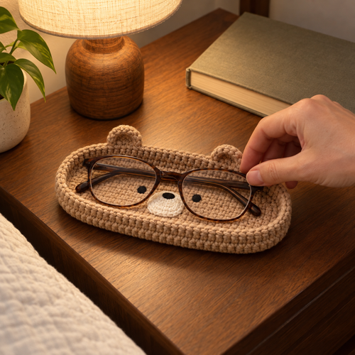 An adult hand places folded reading glasses into a tan bear crochet tray on a bedside table beside a lamp and closed book.