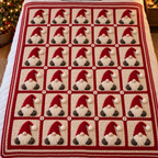 Red-framed cream granny-square blanket with upright red-hat gnomes and striped edging on a made bed beside a Christmas tree.