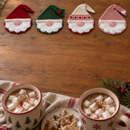 Four flat gnome coasters with red, green, cream and blush hats on a wooden table near cocoa mugs.