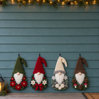 Four floral Christmas gnome door hangers with poinsettias, holly, winter flowers and woodland details hanging on a blue wooden wall.