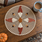 Ecru autumn crochet doily with rust and terracotta gnome hats, a rounded oak leaf and four acorns on a coffee table beside tea and a pumpkin.