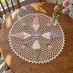 Cream spring crochet doily with pastel gnome hats, a lilac flower and four pink tulips on a kitchen table under a small vase of tulips.