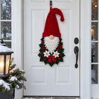 Red-hatted crochet gnome with holly leaves, red berries, white flowers and a red ribbon bow on a white front door beside a snowy lantern.