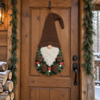 Brown-hatted crochet gnome with pinecones, green sprigs and red-capped mushrooms on a cabin door beside stacked logs.