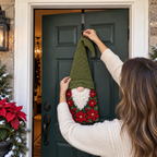 An adult hanging a finished green-hatted poinsettia crochet gnome from an over-door wreath hook.