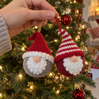 An adult hand holds Classic and Candy Stripe gnome baubles by their hanging loops beside a Christmas tree.
