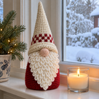 A cream-hatted crochet gnome with a red-and-white band, red body and white loop beard stands on a snowy windowsill beside a candle.