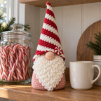 A pink-bodied crochet gnome with a red-and-white spiral hat and peppermint brim detail stands on a kitchen shelf beside a jar of candy canes.