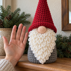 An adult hand beside the classic gnome shows the pink crochet nose, red hat brim and white loop-stitch beard at close range.