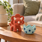 Terracotta crocheted Triceratops with three ivory horns standing beside its empty mint egg with navy spots on a wooden table.