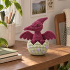 Plum crocheted Pterodactyl with spread scalloped wings in a pale sage egg with lavender spots on a wooden shelf beside notebooks and a pencil cup.