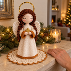 A crocheted Angel holds a gold star on a cream doily, with an adult hand resting beside her on the mantel for scale.