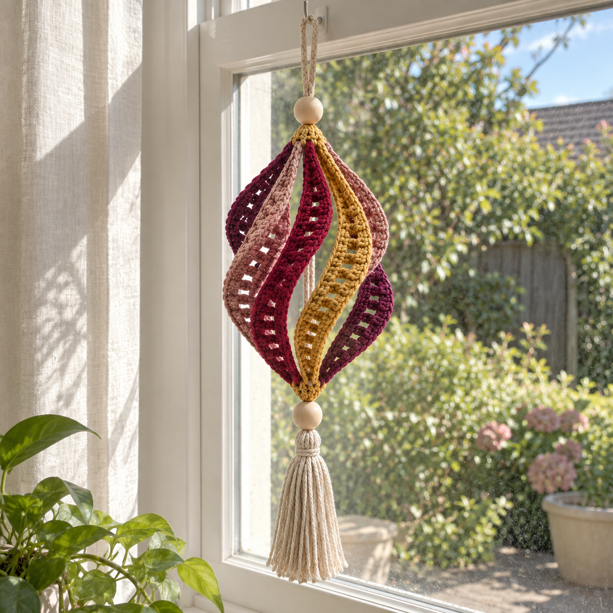A plum, dusty rose, wine and gold Ruby Lantern crochet wind spinner hanging in a bright window beside a linen curtain.