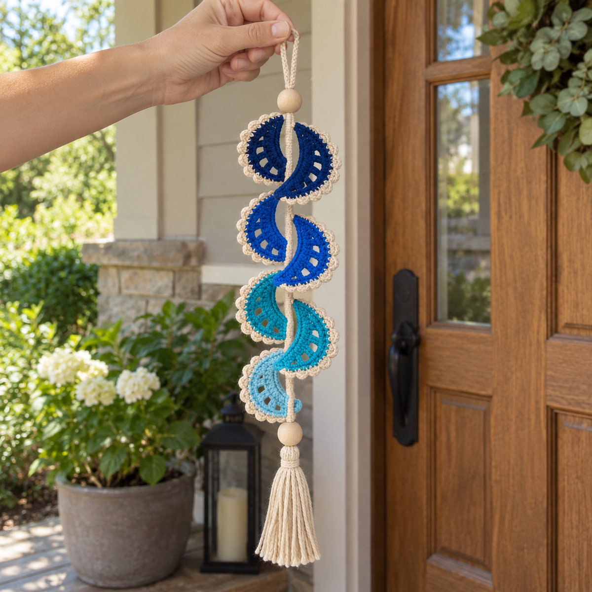 An adult hand holding the oatmeal hanging loop of a Sapphire Crescent crochet wind spinner beside a wooden home door.