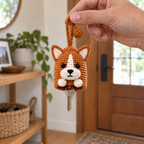 An adult hand holding a Corgi crochet key pouch by its pull loop with a house key below.