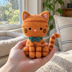 An adult hand holding an orange striped crochet kitten with a teal bow tie beside a sunny window.