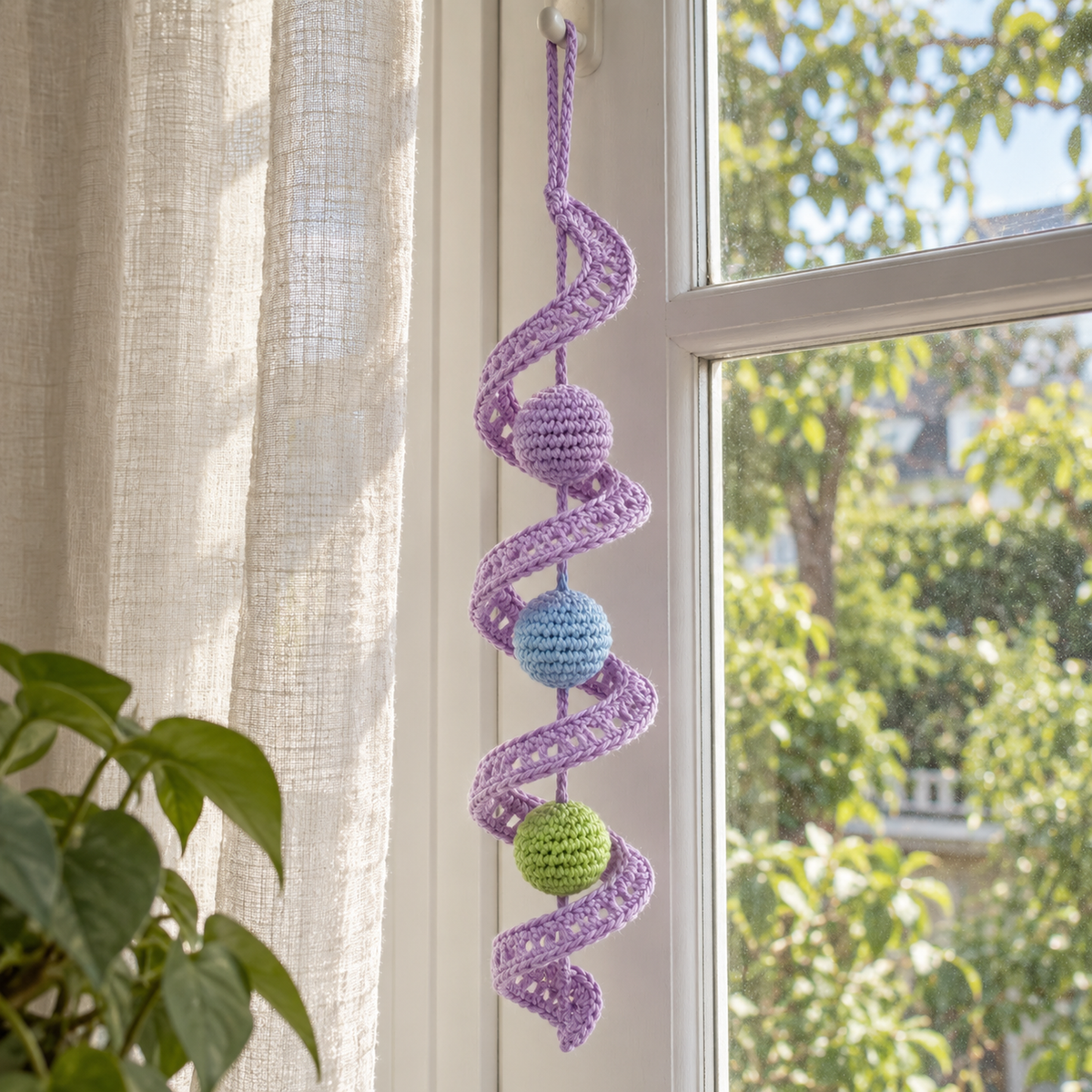 A lilac openwork Lace Twist with lilac, pale blue and green crochet balls hangs beside a sheer curtain in a bright window.