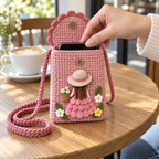 An adult hand slides a smartphone into the open blush pink crochet Flower Basket Lady phone bag on a wooden café table.