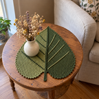 A sage and moss green Heart Linden crochet placemat beneath a vase of dried flowers on a round wooden side table beside an armchair.