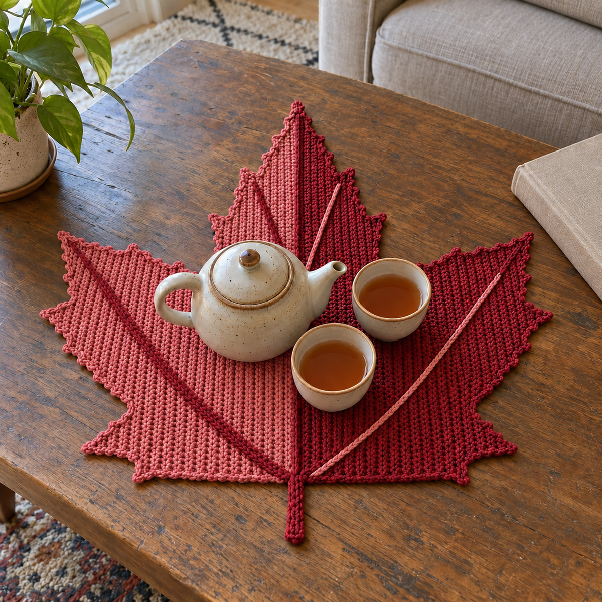 A dusty rose and cranberry Red Maple crochet placemat beneath a teapot and two cups on a wooden coffee table, with a closed book nearby.