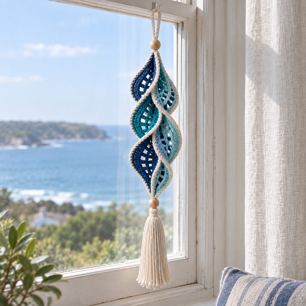 Ocean Blade hangs in a coastal window, with navy, teal and pale aqua crochet leaves, cream edges, wooden beads and a cream tassel.