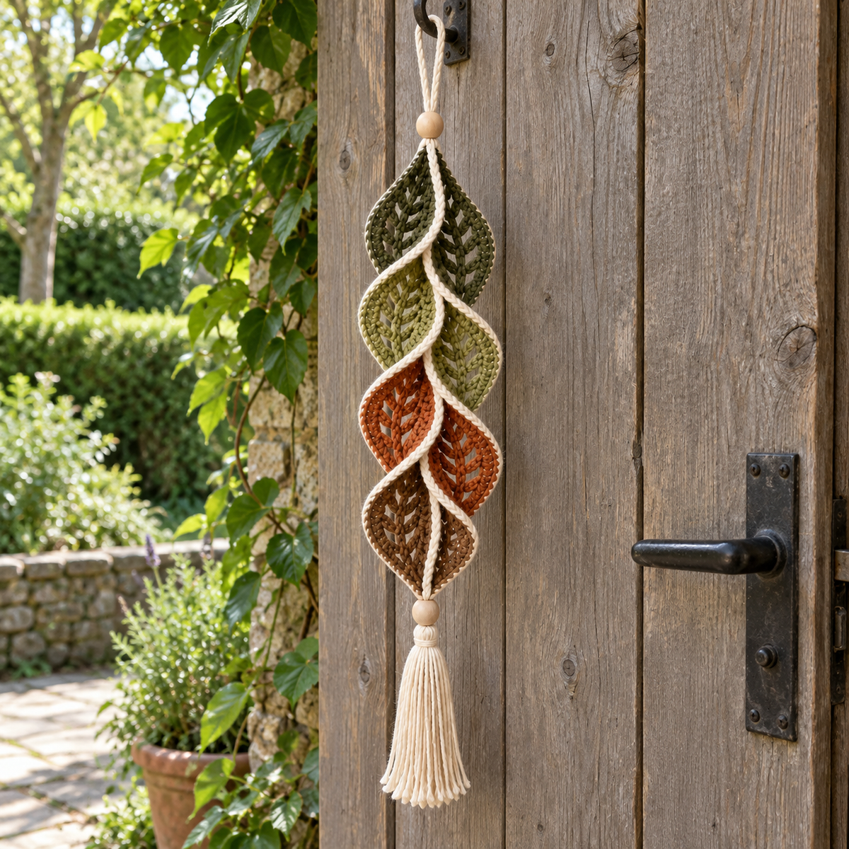 Woodland Vein hangs from a hook by a weathered wooden garden door, with olive green, rust and brown openwork leaves, cream edging and a cream tassel.