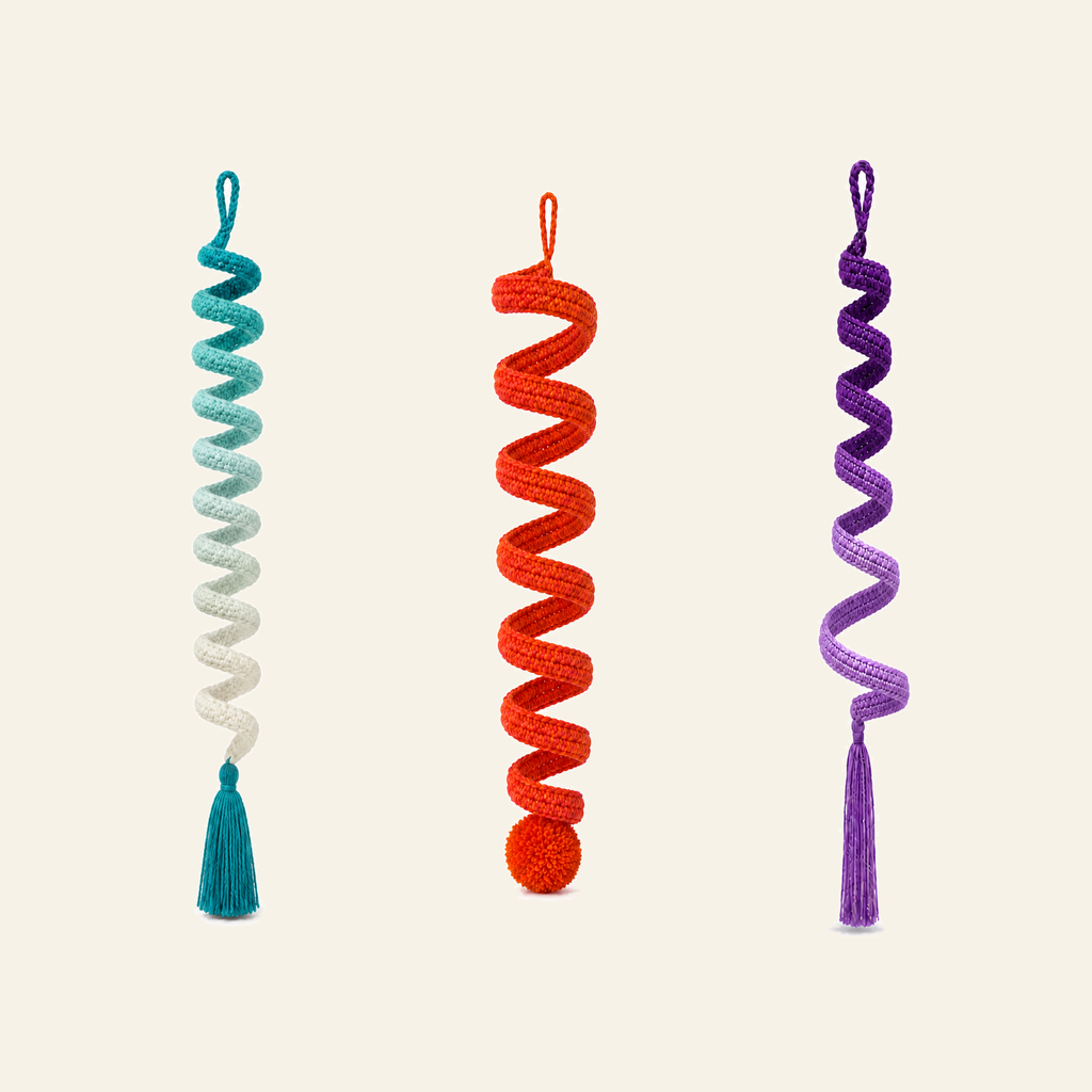 Three crochet spiral wind spinners side by side, teal, sunset orange and violet, each a long corkscrew band with a tassel or pompom at the bottom
