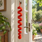 An orange-red crochet spiral wind spinner with a round pompom hanging beside an open garden door.