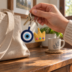 An adult hand lifts a four-colour evil-eye disc and three-point gold crown with pink, aqua and purple embroidered band dots beside a canvas bag.
