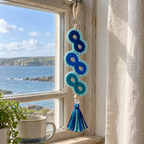 A blue and teal Ocean Infinity crochet ornament hanging in a coastal window.