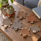Fox, bear, squirrel, hedgehog, raccoon, deer and owl crochet coasters on a living-room coffee table, with a tea mug resting on the fox coaster.