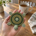 An adult hand holding a green turtle crochet coaster above a wooden table beside a mug of tea.