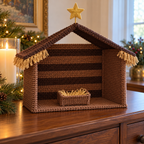 Brown striped crochet stable with a pitched roof, straw-colored yarn fringe, gold star and small loop-filled manger beside a candle.