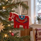 Red Dala horse crochet ornament with a cream mane and tail and colourful folk embroidery hanging on a lit Christmas tree.