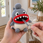 Adult hands hold a grey and white Shark crochet key pouch and pull two keys from its red rolled mouth while the grey ball rests on its head.