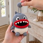 The same Shark crochet key pouch rests in an adult hand while the other hand lifts its grey ball on two chain strands and the keys are hidden inside.