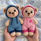 Two adult hands hold blue and pink crochet teddy bears above a soft blue patchwork quilt.