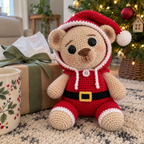 A Santa crochet teddy bear sits beside a wrapped gift and tissue paper near a Christmas tree.
