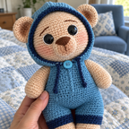 Close view of a blue crochet teddy bear showing its round eyes, embroidered nose, hood trim and neck button, supported by adult fingers.