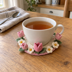 A white teacup of tea on an ivory crochet coaster with pink tulips, ivory flowers and green leaves around its foot on a kitchen table.