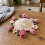 An empty warm-ivory crochet dome with pink tulips, ivory flowers and a soft-pink ruffled flower splayed around its rim on a wooden kitchen table.